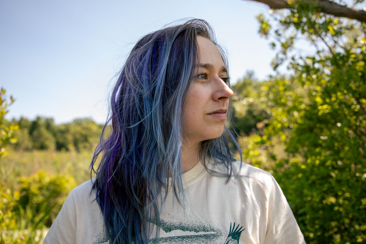 Ashley stands in a sunny marsh, gazing off camera to the viewer's right. Her hair is deep blue, streaked with pale blue and purple. Cattails and button bushes fade off into the distance behind her.
