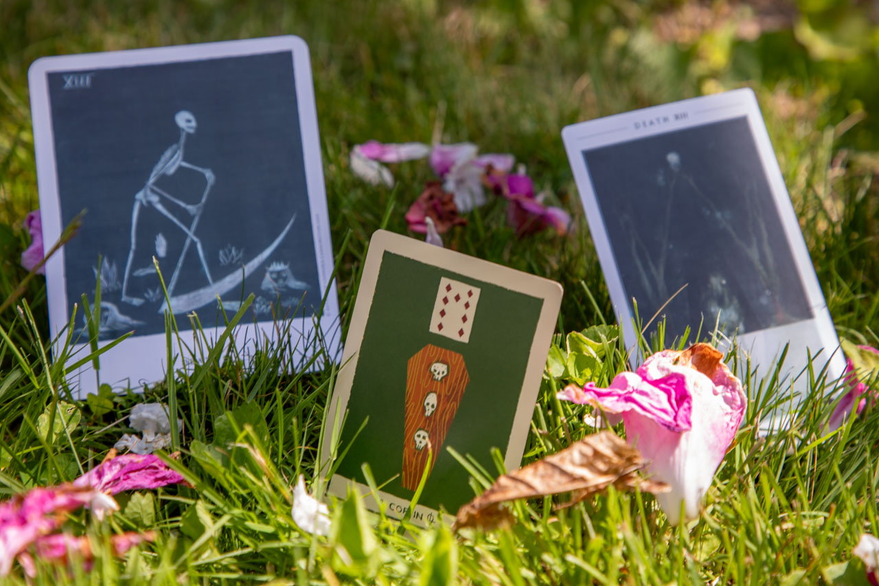 Divination cards arrayed like gravestones in green grass, scattered amongst pink peony petals. In the foreground is a card labeled Coffin, with a brown coffin depicted beneath the 9 of Diamonds. In the background are two renditions of the Tarot’s Death card, one depicting a gangly skeleton reaping with an enormous scythe, the other showing a skeletal horse in moonlit woods.